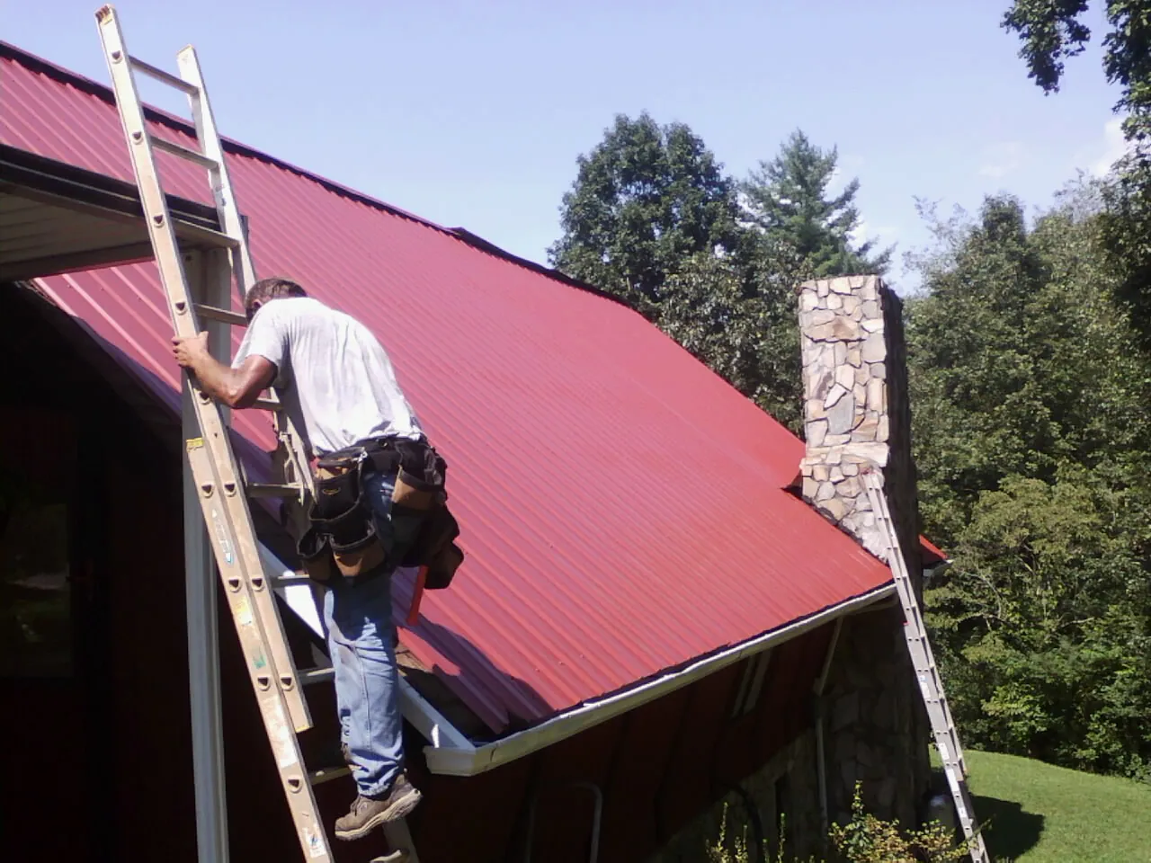 Roofer on a ladder beside a red metal roof and stone chimney — Soffit & Fascia Repair by Roofing Brothers in Granby